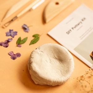 K&ouml;nnte beinhalten: Ein DIY-T&ouml;pferset mit einer wei&szlig;en Tonscheibe, getrockneten Blumen und Werkzeugen auf einer hellorangen Oberfl&auml;che. Das Set enth&auml;lt ein Holz-Formwerkzeug, ein Metallwerkzeug und einen Holz-Schaber. Der Text "DIY Pottery Kit" ist auf einer wei&szlig;en Karte gedruckt.