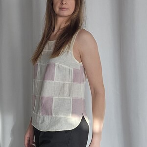 May include: A sleeveless tank top with a patchwork design. The top features a cream-colored base with rectangular patches of light purple. The bottom half of the image shows black pants.