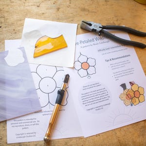 Puede incluir: Un patrón de vitral para una flor silvestre de cinco pétalos con instrucciones y herramientas. El patrón incluye una flor de seis pétalos, un cortador de vidrio y un par de alicates. Las instrucciones incluyen consejos y recomendaciones para cortar el vidrio.