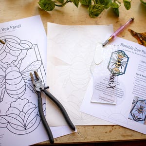 May include: A stained glass bee panel craft kit with a black and white bee design, a clear plastic template, a pair of pliers, and instructions. The instructions include the text "Bumble Bee Panel" and "Difficulty Level: Expert".