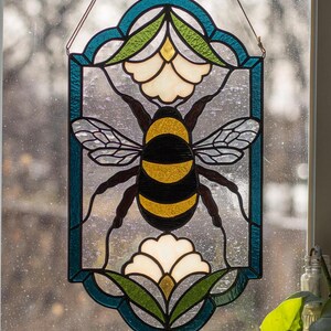 Puede incluir: Una ventana de vitral colgada en un marco de ventana. La ventana presenta una abeja con rayas negras y amarillas, rodeada de flores blancas y hojas verdes. La abeja est&aacute; en el centro de la ventana, con las flores y las hojas que la rodean. La ventana est&aacute; enmarcada en un borde azul.