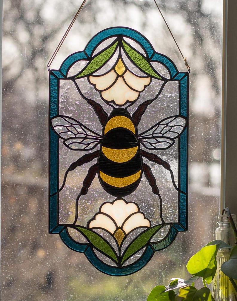 Puede incluir: Una ventana de vitral colgada en un marco de ventana. La ventana presenta una abeja con rayas negras y amarillas, rodeada de flores blancas y hojas verdes. La abeja est&aacute; en el centro de la ventana, con las flores y las hojas que la rodean. La ventana est&aacute; enmarcada en un borde azul.