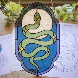 Puede incluir: Un panel de vitral que representa una serpiente verde enroscada alrededor de una luna blanca sobre un fondo azul. La serpiente tiene estrellas amarillas en su cuerpo. El panel está enmarcado con un borde gris oscuro.