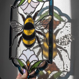 Puede incluir: Un panel de vitral que representa una abeja con rayas negras y amarillas, posada sobre una flor blanca con hojas verdes. El panel est&aacute; enmarcado en metal negro y tiene un fondo azul y verde.