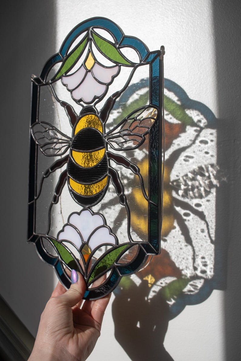 Puede incluir: Un panel de vitral que representa una abeja con rayas negras y amarillas, posada sobre una flor blanca con hojas verdes. El panel est&aacute; enmarcado en metal negro y tiene un fondo azul y verde.