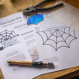 May include: A close-up of a glass spider web pattern with instructions for creating a stained glass project. The pattern includes a dotted grey line indicating zinc came placement and a dotted circle indicating hook placement. The text on the page reads "Glass Spider Web, A pattern by Rosie Linebaugh, Difficulty Level: Intermediate, Lots of concave curves with sharp pri, Tips & Recommend: Zinc came is necessary along the edge of this project. I use, Cut the concave curves! Preserve the pointy tip of glass, Dotted circles indicate hook placement, Dotted grey line indicates zinc came placement, Please do not reproduce, share or publish this pattern. This pattern is for both personal and commercial use, so you are welcome to sell this pattern. If posting it online, be sure to tag Rosie Linebaugh/ Linebaugh Studios LLC, Copyright is retained by Linebaugh Studios LLC".