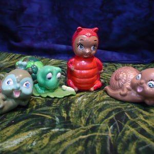 May include: A collection of whimsical ceramic figurines, including snails, a turtle, and a ladybug. The snails have brown and teal bodies with red and brown shells. The ladybug is red with a smiling face. The turtle is green.