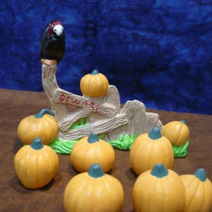 May include: A Halloween-themed ceramic decoration featuring a black bird perched atop a fence with the word "BEWARE" and several orange pumpkins with green stems. The pumpkins are scattered around the fence.