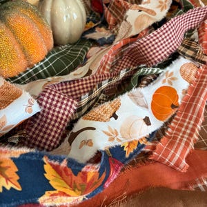 Könnte beinhalten: Eine Ansammlung von Stoffstreifen mit herbstlichen Mustern wie orangefarbenen Kürbissen, Eicheln und Herbstblättern, durchsetzt mit einfarbigen und karierten Bändern in Rot-, Grün-, Braun- und Marineblautönen sowie zwei kleinen dekorativen Kürbissen.