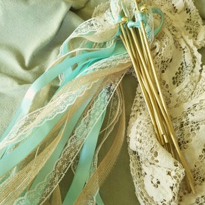 May include: A set of wedding ceremony ribbon wands with light blue satin ribbon, burlap ribbon, and white lace. The wands have gold sticks with bells at the top.