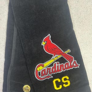 Puede incluir: Toalla negra con el logotipo de los Cardenales de San Luis bordado en rojo, amarillo y blanco. El logotipo presenta un cardenal posado en un bate de béisbol, con la palabra "Cardinals" debajo. Las iniciales "CS" están bordadas en amarillo.