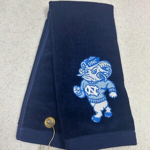 May include: Navy blue towel with a light blue and white embroidered ram mascot and "UNC" logo. The towel has a dark blue border and a metal hook. The towel is folded in thirds.
