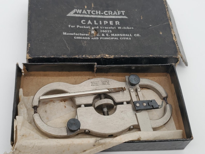 May include: A silver-toned metal Watch-Craft caliper for pocket and bracelet watches, featuring a central adjustment wheel and black knobs, housed in a black cardboard box with white text reading Watch-Craft Caliper and C. & E. Marshall Co.