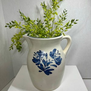 Könnte beinhalten: Eine weiße Keramikkanne mit blauem Blumendesign. Die Kanne hat einen Henkel und einen Ausguss. Sie ist mit grünen, blättrigen Stielen gefüllt.