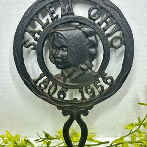 May include: Black cast iron hot pad with a raised design of a woman's face and the text "Salem Ohio 1806 1956".