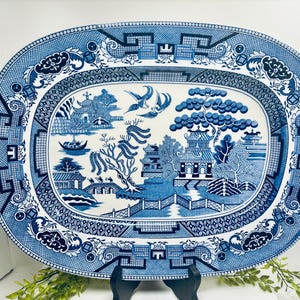 May include: An oval ceramic platter featuring a blue and white willow pattern. The design depicts a landscape with pagodas, a bridge, a willow tree, and two birds in flight. The wide border is decorated with intricate geometric and floral motifs.