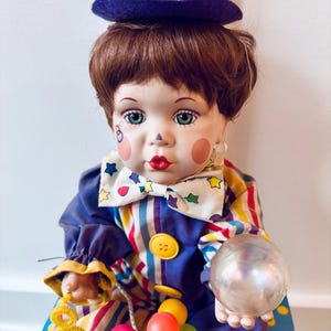 May include: A porcelain clown doll wearing a blue and yellow striped outfit, a purple hat with colorful stripes, and a bow tie. The doll is holding a clear glass ball and is surrounded by colorful balloons and a yellow spring.