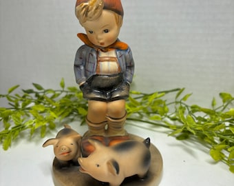 May include: A figurine of a boy in a blue and white shirt and orange hat standing with two small pig figurines. The boy is looking down at the pigs.