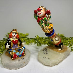 May include: Two clown figurines. One clown is seated with a gold hat, black jacket, and red bow tie. The other clown is inside a cannon. Both are on stone bases. The cannon has the text "Best Wishes" and "Ray Jun 96".