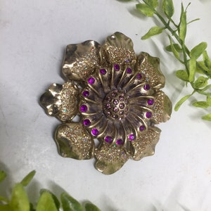 May include: A gold flower brooch with purple glitter and rhinestones.