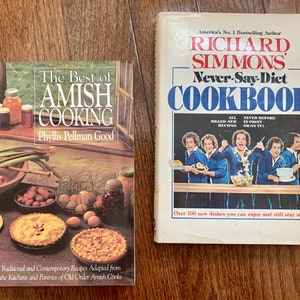 May include: Two cookbooks, one titled "The Best of Amish Cooking" by Phyllis Pellman Good, featuring a photo of a kitchen scene with pies and a bowl of fruit, and the other titled "Richard Simmons' Never-Say-Diet Cookbook" featuring a photo of Richard Simmons and other people holding food.