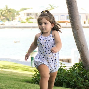 May include: A young girl in a white and blue polka dot romper runs on a grassy lawn. The romper has a halter neck and a gathered skirt.