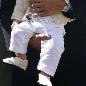 May include: A baby wearing a white knitted cardigan and white pants is being held by an adult. The baby is wearing a white crocheted scarf around their neck. The adult is wearing a dark blue suit jacket.
