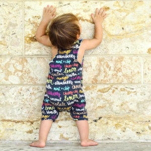 May include: A child wearing a black and white jumpsuit with colorful text that reads "mango", "strawberry", "mint + chip", "cherry chip", "coconut", "lemon", "vanilla", and more. The child is standing in front of a stone wall.