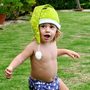 May include: A young child wearing a green and white sleep cap with a white pom-pom on top. The child is wearing a blue and white patterned diaper and is standing on a green grassy lawn.