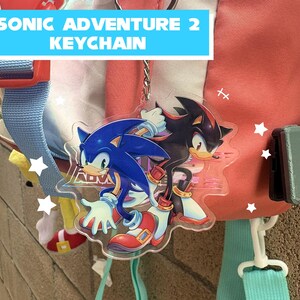 May include: A clear acrylic keychain featuring Sonic the Hedgehog and Shadow the Hedgehog from Sonic Adventure 2. The keychain has a silver chain and the text "SONIC ADVENTURE 2 KEYCHAIN" in a blue banner.