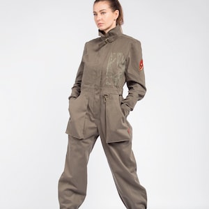 Op de afbeelding: Een vrouw draagt een groene utility jumpsuit met een patch op de mouw. Ze staat met haar handen in haar zakken en draagt groene laarzen.