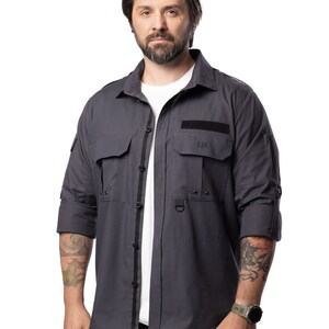 May include: A charcoal gray button-down shirt with two chest pockets and a black accent stripe down the front. The shirt has roll-up sleeves and the letters "UA" are visible on one pocket.