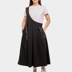 Puede incluir: Una mujer con una camiseta blanca y una falda negra con un dise&ntilde;o asim&eacute;trico &uacute;nico. La falda tiene pliegues y una silueta larga y fluida. Lleva zapatillas blancas.