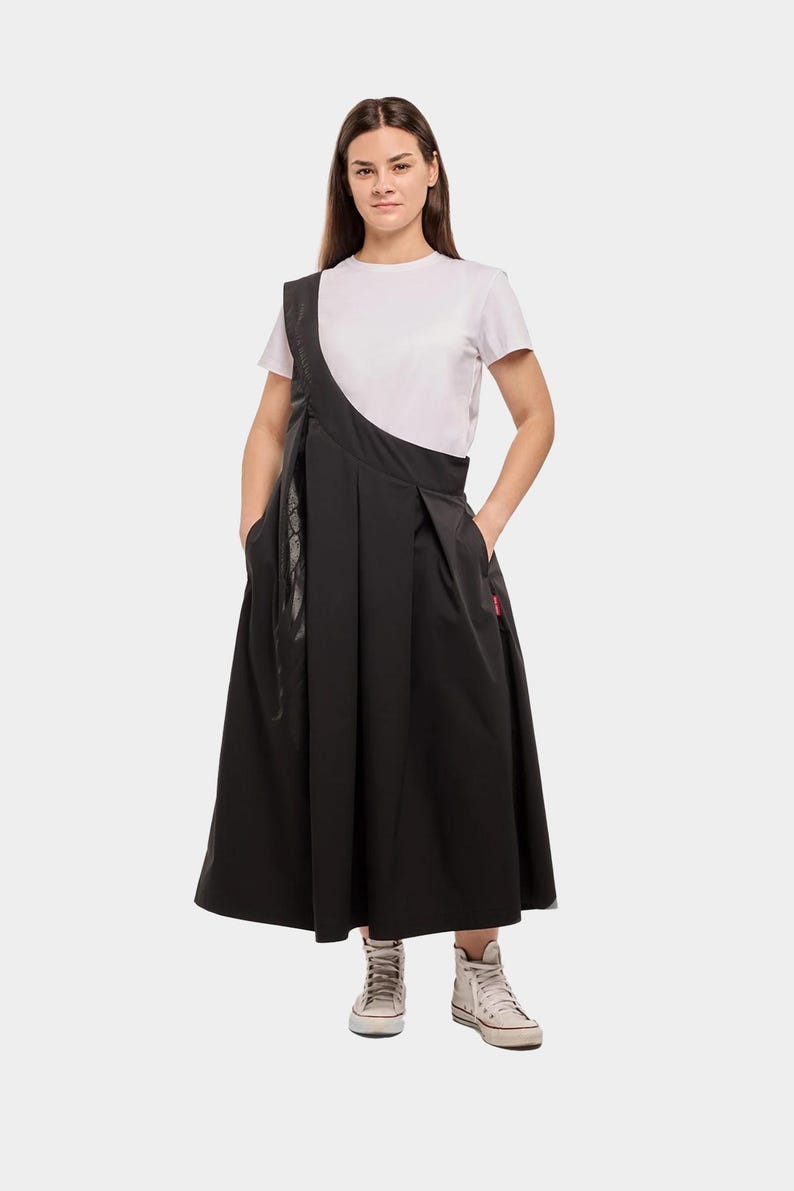 Puede incluir: Una mujer con una camiseta blanca y una falda negra con un dise&ntilde;o asim&eacute;trico &uacute;nico. La falda tiene pliegues y una silueta larga y fluida. Lleva zapatillas blancas.