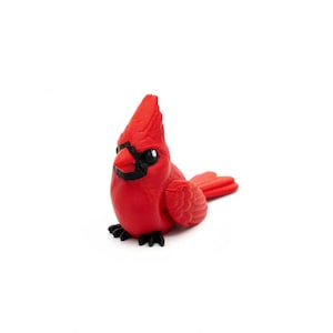 May include: A small, red cardinal figurine with black accents around the eyes and beak. The bird is perched on a surface, showcasing its detailed feathers and a distinctive crest. The figurine is a decorative item.