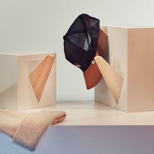 Könnte beinhalten: Eine schwarze Trucker-Cap mit braunem Schirm und Mesh-Rückseite wird auf einem hölzernen Akzentstück präsentiert. Eine gefaltete beige Mütze liegt auf einer weißen Oberfläche. Die Szene zeigt geometrische Formen und eine minimalistische Ästhetik.