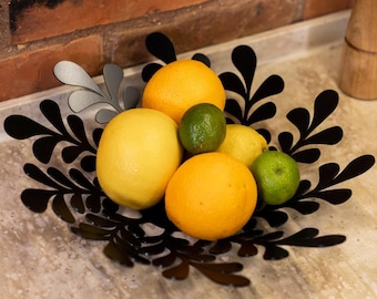 May include: A black metal bowl with a leafy design holds a variety of citrus fruits, including lemons, limes, and oranges.