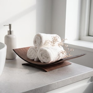 Puede incluir: Un lavabo de cerámica blanca, un dispensador de jabón blanco con bomba plateada y una bandeja de madera marrón oscura que contiene tres toallas blancas enrolladas y una pequeña rama de flores secas, sobre una encimera de color gris claro.