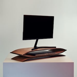 Peut inclure: Un support d'écran en bois foncé présentant un design incurvé à deux niveaux avec une finition lisse. Le support soutient un écran d'ordinateur noir sur l'étagère supérieure, tandis qu'un ordinateur portable fin et fermé repose sur l'étagère inférieure.