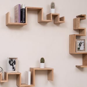 Op de afbeelding: Een set lichtbruine houten wandplanken met een zigzagontwerp. De planken zijn gerangschikt in drie afzonderlijke secties en bevatten twee kleine cactussen in potten, twee boeken en twee rechthoekige fotolijsten met zwart-witfoto's.