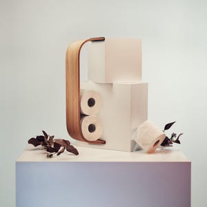Puede incluir: Un moderno portarrollos de papel higiénico con un asa de madera curvada y un diseño de bloques blancos. Se ven dos rollos de papel higiénico en el soporte. Hojas secas decorativas se colocan alrededor.
