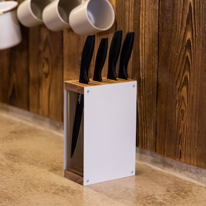 May include: A white knife block with four black kitchen knives. The block is made of wood and has a white acrylic front panel. The knives are arranged in the block with their handles facing up.
