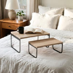 May include: Two rectangular bed trays with light-toned wooden tops and black metal U-shaped legs. The larger tray holds an open book and a black ceramic mug. The trays are positioned on a white textured bedspread with pillows in the background.