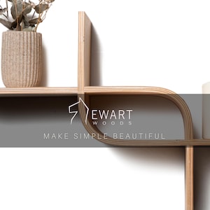 May include: A modern, light-colored wooden wall shelf with a unique, geometric design. The shelf features curved and straight lines, creating an open, airy aesthetic. A beige textured vase with dried plants sits on the shelf. The text "EWART WOODS MAKE SIMPLE BEAUTIFUL" is visible.