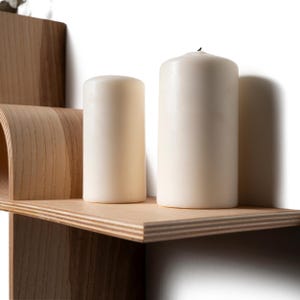 May include: A wooden shelf with a curved design and two ivory-colored pillar candles. The candles are cylindrical and vary slightly in height. The shelf is made of light-colored wood and has a clean, modern aesthetic.