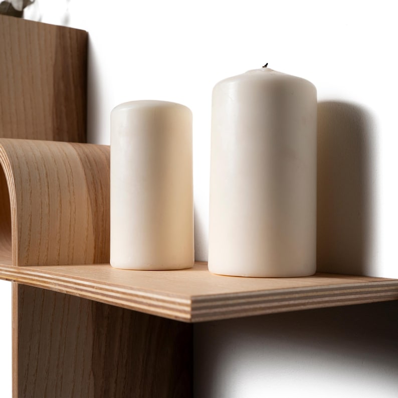 May include: A wooden shelf with a curved design and two ivory-colored pillar candles. The candles are cylindrical and vary slightly in height. The shelf is made of light-colored wood and has a clean, modern aesthetic.