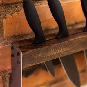 Close-up of a stylish knife holder with a dark wooden block and visible black screws, holding black-handled knives against a rustic red brick kitchen wall.