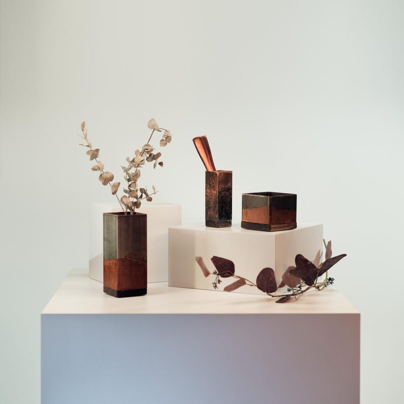 May include: A still life arrangement featuring three geometric copper-toned containers. One holds dried branches, another has a copper utensil, and the third is a small square box. The objects are displayed on white pedestals, with a neutral background.