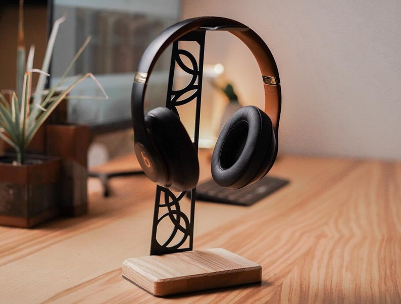 Handcrafted headphone stand with a smooth birch wood base and a black metal frame featuring intricate cutout patterns, holding over-ear headphones on a wooden desk in a modern workspace.