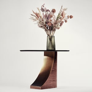 May include: A modern side table with a black glass top and a curved, bronze-toned metal support. The base is made of wood. A vase of dried flowers in shades of brown, beige, and purple sits on the table.